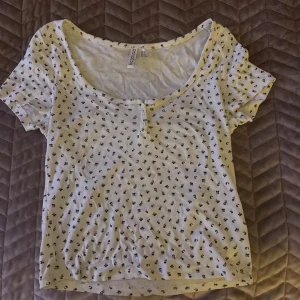Vit blommig topp från H&M Divided - Supersöt vit topp från H&M Divided med små svarta blommor över hela. Rundad halsringning, korta ärmar och knappar framtill. Mjuk och skön bomullskvalitet, perfekt för vår och sommar. Enkel att matcha med jeans eller kjol för en chill look. Jag har bara använt den 2 gånger och gillar inte hur den sitter på mig så därför säljer jag den. Pris kan diskuteras. 