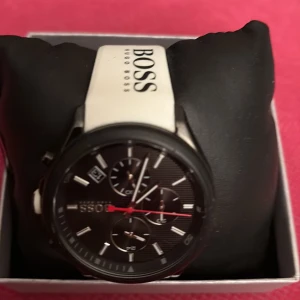 Svart kronografklocka Hugo Boss - Snygg kronografklocka från Hugo Boss med svart urtavla, vita index och detaljer i rött. Klockan har en vit rem med BOSS-logga och flera små urtavlor för extra funktioner. Rund boett i svart metall och sportig design som sticker ut.