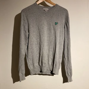 Grå stickad tröja från Lyle & Scott - Snygg grå stickad tröja från Lyle & Scott med klassisk v-ringning och broderad logga på bröstet. Tröjan har ribbade muddar och är långärmad, perfekt för lager-på-lager under hösten och vintern. Står M men sitter mer som S. Hör av dig vid minsta fundering!