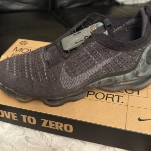 Nike Vapormax Flyknit svart sneaker - Nike Vapormax Flyknit sneakers i mörkgrått med stickad ovandel och genomskinlig, bubblig sula. Snyggt mönstrad innersula och diskret Nike-logga på sidan. Skon har snörning med smart lås och är tillverkad i flyknit-material för en lätt och flexibel känsla.