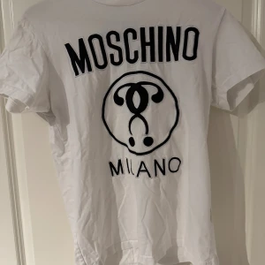 Vit Moschino Milano t-shirt - Snygg vit t-shirt från Moschino med stort svart Moschino Milano-tryck på framsidan. Klassisk rund hals och korta ärmar. Materialet är mjuk bomull som känns skönt mot huden. Perfekt för dig som gillar statement-plagg och exklusiva märken.