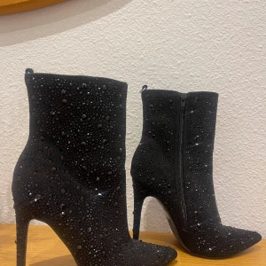 Svarta glittriga stövletter med klack - Säljer ett par svarta stövletter med spetsig tå och hög klack. Skorna är täckta av svarta glittriga stenar som ger en riktigt lyxig och festlig känsla. Materialet är mockaliknande och de har dragkedja på sidan för enkel påtagning.