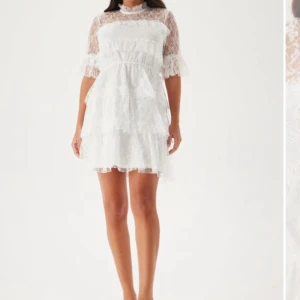 Vit spetsklänning med volanger - Superfin vit kortklänning i spets med volanger och halvtransparent överdel. Klänningen har högt halslinje, korta ärmar med volangkant och flera lager i kjolen för extra fluff. Perfekt konfirmation/student klänning. Bara använd en gång (konfirmation)