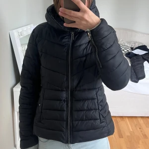 Svart pufferjacka från Etirel, strl 34 - Säljer en svart pufferjacka från Etirel i storlek 34. Jackan har huva, dragkedja framtill och quiltad design. Perfekt för kyliga dagar och har en snygg, enkel look som passar till det mesta. En knapp på ena fickan har lossnat men stör inte alls (se bild )😊🎀