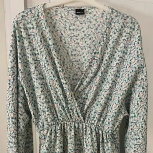 Blommig omlottklänning Gina - Supersöt kortklänning från Gina tricot  med blommigt mönster i ljusa toner av rosa, blått och vitt. Klänningen har omlott-v-ringning, långärmade puffärmar och resår i midjan för en skön passform. Perfekt för vår och sommar!