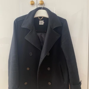 Svart skepparkavaj  - Klassisk svart skepparkavaj med dubbelknäppning och bred krage. Jackan är tillverkad i ullblandning och har två snedställda fickor framtill samt slits bak. 