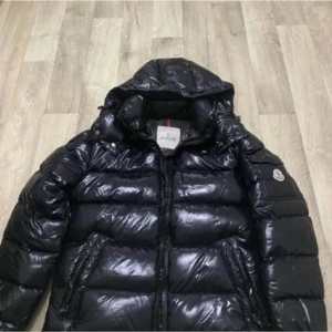 Svart dunjacka från Moncler med huva - Säljer en svart, glansig dunjacka från Moncler med huva och dragkedja. Jackan har klassisk pufferstil, två fickor med dragkedja och Moncler-logga på ärmen. Perfekt för kalla dagar och riktigt snygg streetstil.