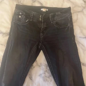 Mörkgrå jeansbyxor från Gina Tricot - Mörkgråa jeansbyxor från Gina Tricot med vita kontrastsömmar och dubbla knappar i midjan. Klassisk femficksmodell och raka ben. Snygga till sneakers eller boots och passar perfekt till en avslappnad streetstil.