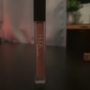 Huda Beauty Liquid Matte Bombshell - Säljer ett Huda Beauty Liquid Matte läppstift i färgen Bombshell. Det är en flytande formula med matt finish i en snygg nude-rosa ton. Kommer i en stilren, frostad tub med svart lock. Perfekt för dig som gillar trendiga och hållbara läppfärger. Aldrig använd, öppnad för att se färg 