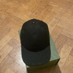 Svart keps från Gucci med GG-mönster - Säljer en svart keps från Gucci med diskret GG-monogram i tyget. Kepsen har klassisk form, böjd skärm och justerbart spänne bak i skinn. Sidorna pryds av Guccis ikoniska gröna och röda band. Perfekt för dig som vill ha en lyxig och stilren accessoar. Om du har någon fråga är det bara att skriva! Priset kan diskuteras vid SNABB affär!😁