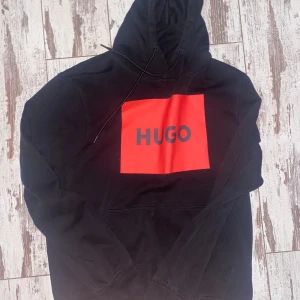 Svart hoodie från Hugo med röd logga - Svart hoodie från Hugo med stor röd fyrkant och HUGO-tryck på bröstet. Klassisk huva med snörning och känguruficka framtill. Mjuk bomullskänsla och långärmad modell, perfekt för en chill och stilren look.