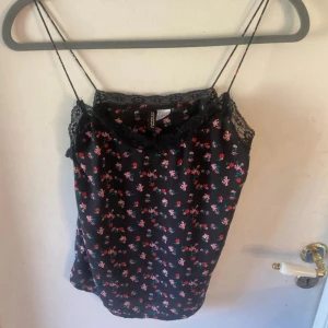 Svart blommig linnetopp med spets - Svart linnetopp från Cropp med tunna axelband och blommigt mönster i rosa, rött och grönt. Toppen har dekorativ spets längs halsringningen och är ärmlös med en loose passform. Perfekt för sommarens alla häng!