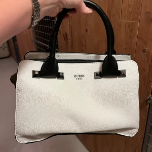 Vit och svart handväska från Guess - Snygg handväska från Guess i vit, grå och svart med stilrena detaljer i silver. Väskan har två rejäla handtag, strukturerad form och flera fack inuti för smart förvaring. Materialet är skinnimitation med en lyxig känsla och coola metallnitar framtill.