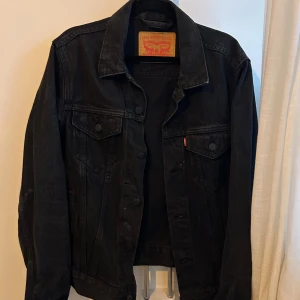 Svart jeansjacka från Levi's - Snygg svart jeansjacka från Levi's i klassisk modell med krage och knappar framtill. Jackan har två bröstfickor med lock och knapp, samt raka sömmar och en cool vintage-vibe. Perfekt för dig som gillar streetstyle och tidlös design.