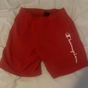 Röda Champion shorts med tryck - Snygga röda shorts från Champion med vit logotyp längs ena benet. Modellen har elastisk midja och är tillverkad i mjukt material som passar perfekt för chill eller sport. Klassisk och enkel design med fickor på sidorna.