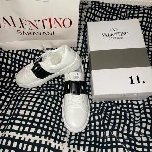 Valentino Garavani Rockstud sneakers - Snygga vita Valentino Garavani Rockstud sneakers med svart läderdetalj och ikoniska nitar över vristen. Skorna har rund tå, platt sula och klassisk snörning. Lyxig känsla med exklusiv design och cool kontrast mellan vitt och svart. Perfekta för dig som vill sticka ut.