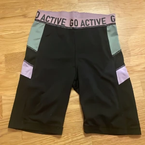 GO ACTIVE svarta träningstights-shorts - Svarta träningstights-shorts från GO ACTIVE med bred lila resår i midjan och text. Har grå och lila detaljer på sidorna. Sitter tight och är perfekta för gym eller löpning. Gjorda i stretchigt syntetmaterial som andas. Lite noppriga. 
