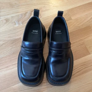 Svarta loafers från Ami - Snygga svarta loafers från Ami Alexandre Mattiussi med chunky platt sula och klassisk penny-detalj framtill. Skorna är tillverkade i blankt skinn och har en fyrkantig tå samt en liten metallhjärta-detalj bak på hälen. Perfekta för en stilren och modern look. Köpta på Vestiaire Collective. Digitalt inspekterade av deras team. Fick inte med kvittot av säljaren eller dustbag därav priset. Endast använda två gånger. Kan gå ner i pris vid snabb affär 🥳