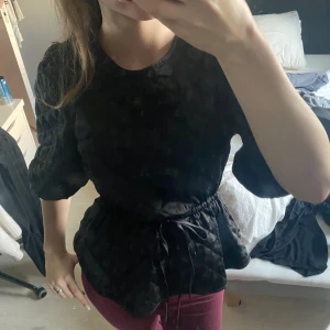 Svart quiltad blus med knyt i midjan - Säljer en svart quiltad blus med puffiga trekvartsärmar och rund halsringning. Blusen har ett snyggt knytband i midjan. Varm och skön blus.
