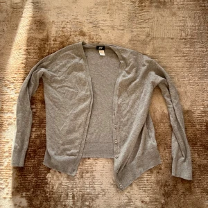 Grå kofta från H&M med knappar - En enkel och stilren grå kofta från H&M med v-ringning och knäppning framtill. Perfekt att slänga över en topp eller t-shirt för en chill look.