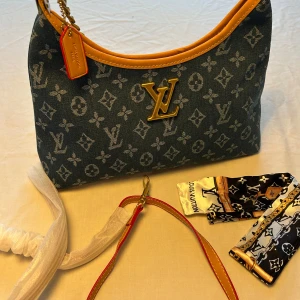 Louis Vuitton denim axelväska - Snygg axelväska Vuitton i blå denim med klassiskt monogrammönster och detaljer i ljusbrunt skinn. Väskan har guldfärgade metalldetaljer, kedja och LV-logga framtill. Medföljer extra axelrem och scarf med LV-tryck.