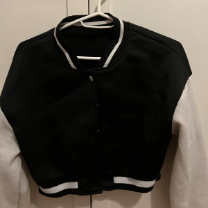 Svartvit croppad bomberjacka - Snygg croppad bomberjacka i svart och vitt med klassisk collegekrage och tryckknappar. Jackan har vita ärmar och svarta muddar med vita ränder. Perfekt för en sportig och trendig look.