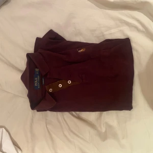 Vinröd pikétröja från Polo Ralph Lauren - Säljer en vinröd långärmad pikétröja från Polo Ralph Lauren i classic fit. Tröjan har bruna detaljer vid knäppningen, tre knappar och den klassiska broderade loggan på bröstet. Perfekt för dig som gillar stilren och tidlös design.