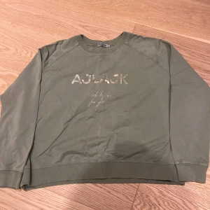 Olivgrön sweatshirt från Ajlajk M - Olivgrön sweatshirt från Ajlajk med metallictryck framtill. Tröjan har rund halsringning, långa ärmar och ribbade muddar vid ärmslut och nederkant. Perfekt för en chill och avslappnad stil.
