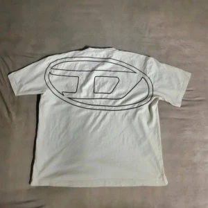Vit t-shirt med stor grafisk logga - Vit t-shirt från Diesel med en stor svart embroidered logga tryckt på ryggen och en mindre logga framtill. Klassisk rund hals och korta ärmar. Boxy fit. Skön passform och tillverkad i mjuk bomull, perfekt för dig som gillar streetwear och skate. Köpt i butik för 100£ i italien. 