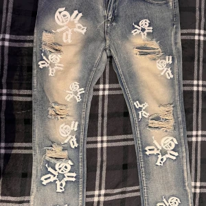 Blå ripped jeans med tryck, XXL - Säljer ett par blå jeans med slitna detaljer och vita tryck i gotisk stil över hela byxorna. Jeansen har en cool, streetig vibe och är tillverkade i 85% bomull och 15% polyester. Perfekta för dig som vill sticka ut med din outfit.