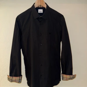 Svart Burberry skjorta - Säljer en svart skjorta från Burberry i bomull. Den är strl M och sitter som M ev L. Litet svart Burberry märke på bröstet och Burberrys signatur rutmönster i krage och manschetter. Galet snygg skjorta som kan användas både casual och festligt. Nypris ligger på 6000kr. 