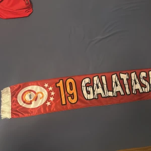 Galatasaray halsduk i rött och orange - Supporterhalsduk för Galatasaray med klubbens logga och årtal 1905. Ena sidan är röd med vita och gula detaljer, andra sidan är orange med röda ränder och vita bokstäver. Halsduken har vita fransar i ändarna och är tillverkad i glansigt syntetmaterial.