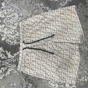 Fendi beige mönstrade shorts - Snygga shorts från Fendi med beige och vitt FF-mönster över hela tyget. De har elastisk midja med svart snörning och innerfoder i mesh. Perfekta för dig som vill sticka ut med lyxig streetstyle.