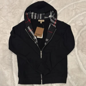 Svart hoodie från Burberry med dragkedja - Svart hoodie från Burberry med klassiskt rutigt foder i huvan och broderad logga på bröstet. Tröjan har hel dragkedja framtill, långa ärmar och huva. Perfekt för dig som vill ha en stilren och lyxig look med ikoniska Burberry-detaljer. Nypris 8000kr. Mitt pris 750kr. Hör av dig för mer information 😁