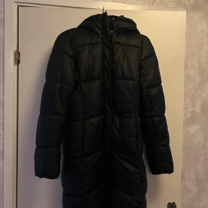 Svart lång pufferjacka med huva - Säljer en svart lång pufferjacka med huva och dragkedja framtill. Jackan har quiltad design och är perfekt för kalla dagar. Den har långa ärmar och en oversized passform. Missfärgad krage av smink