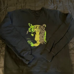 Svart sweatshirt Tiger of Sweden - Svart sweatshirt från Tiger of Sweden med coolt tigertryck i grönt och gult på bröstet. Tröjan är i 100% bomull och har långa ärmar samt ribbade muddar vid ärmslut och nederkant. Perfekt för dig som vill sticka ut med en unik detalj.