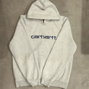 Carhartt hoodie  - Snygg ljusgrå hoodie från Carhartt med broderad mörkblå logga på bröstet. Klassisk modell med huva och ribbade muddar. Perfekt för dig som gillar streetwear och enkel stil.