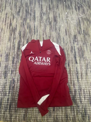 Storlek M PSG zip tröja  - Den ikoniska PSG-loggan och klubbens färger ger tröjan en modern och professionell look – perfekt för dig som vill representera Paris Saint-Germain både på och utanför planen. Oavsett om du använder den till träning, uppvärmning eller som sportig vardagsstil är detta ett plagg som kombinerar funktion och klubbkänsla på bästa sätt.