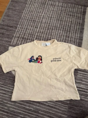 Beige Disney Lilo & Stitch t-shirt Zara - Snygg beige t-shirt från Zara med Lilo & Stitch-tryck och texten 'galactic good time' på bröstet. Croppad modell med rund hals och korta ärmar. Perfekt för dig som gillar Disney och vill ha en chill och unik stil.