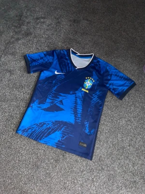 Blå Brasil Nike fotbollströja - Säljer en blå Brasil Nike fotbollströja med grafiskt mönster i olika blå toner. Tröjan har v-ringad krage med vit och marinblå kant, samt Brasil-logga med fem stjärnor på bröstet. Materialet är lätt polyester och perfekt för fotboll eller träning.