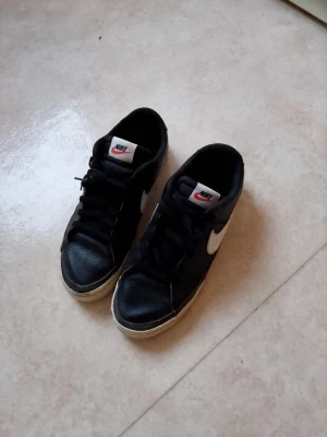 Svarta Nike sneakers med vit swoosh - Klassiska svarta Nike sneakers med vit swoosh på sidorna och vit sula. Skorna har snörning och rund tå, samt Nike-logga på plösen och hälen. Perfekta för dig som gillar en clean och tidlös stil.