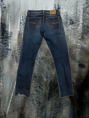 Mörkblå jeans från Nudie Jeans - Snygga mörkblå jeans från Nudie Jeans med klassiska orange sömmar och vågig brodyr på bakfickorna. Jeansen har slim passform, normal midja och är tillverkade i slitstarkt denim. Perfekta för dig som gillar stilrena och tidlösa jeans med coola detaljer. W27 L32 men klippte längst ner så de passar runt 160cm