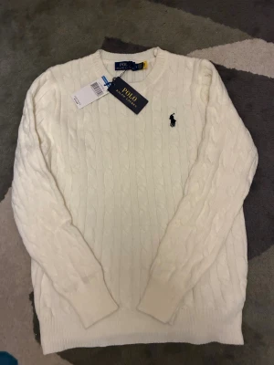 Vit kabelstickad tröja Polo Ralph Lauren - Snygg vit kabelstickad tröja från Polo Ralph Lauren med klassisk rund hals och broderad marinblå logga på bröstet. Tröjan har långa ärmar och ribbade muddar, perfekt för dig som gillar stilren och tidlös design.