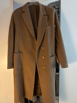 Beige kappa från Tiger of Sweden - Beige kappa från Tiger of Sweden med  krage och dubbelknäppning. Jackan har stora fickor framtill och är helfodrad med slät insida. 