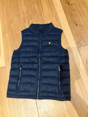 Marinblå dunväst från Lyle & Scott - Snygg marinblå dunväst från Lyle & Scott med gul logga på bröstet. Västen har quiltad design, två fickor med dragkedja och hög krage. Perfekt att slänga över en hoodie för extra värme. Klassisk och stilren look.