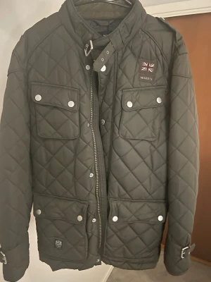 Quiltad grön jacka från Morris, L - Snygg quiltad jacka från Morris i mörkgrönt med fyra stora fickor framtill, silverfärgade knappar och dragkedja. Jackan har hög krage med spänne och coolt brittiskt märke på bröstet. Perfekt för dig som vill ha en stilren och klassisk look.