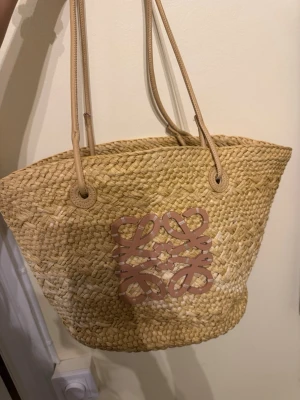 Strandväska  - Snygg stråväska  med klassisk beige färg och långa handtag. Framsidan har loggan i ljusbrunt som ger en lyxig touch. Perfekt storlek för att få plats med allt du behöver och passar till sommarens alla äventyr.