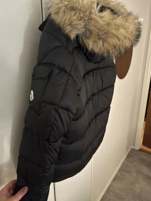 Svart Moncler dunjacka med päls - Säljer en svart Moncler dunjacka oanvänd med stor huva och pälsdetalj. Jackan har dragkedja framtill, två fickor med dragkedja och klassisk Moncler-logga på ärmen. Puffig modell som håller dig varm hela vintern. Perfekt för dig som vill ha både stil och funktion.