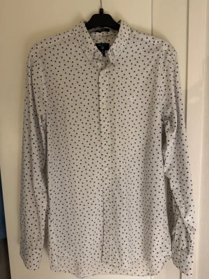 Vit snöflinge-mönstrad skjorta från GANT - Vit skjorta från GANT i storlek M med blått snöflinge mönster och diskret rutmönster. Klassisk krage, långärmad och knäppning framtill. Broderad GANT-logga på bröstet. Skjortan har en normal passform och är tillverkad i mjuk bomull. Ej använd utan prislapp. 