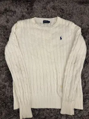 Snygg vit kabelstickad tröja från Polo Ralph Lauren i storlek S. Klassisk design med rund hals och ribbade muddar. Tröjan har det ikoniska blå broderade Polo-logot på bröstet och är långärmad. Perfekt för dig som gillar stilrena och tidlösa plagg.